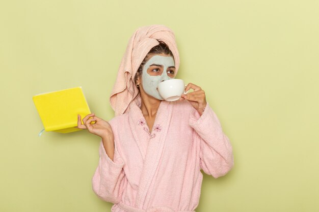 Vista frontal mujer joven en bata de baño rosa tomando café y leyendo el cuaderno sobre la superficie verde