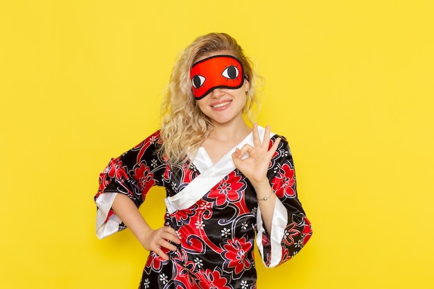 Vista frontal mujer joven en bata de noche y con máscara para los ojos preparándose para dormir posando y sonriendo en la pared amarilla dormir noche de color de oscuridad femenina