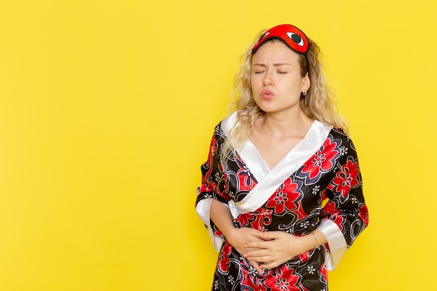 Foto gratuita vista frontal mujer joven en bata de noche y con máscara para los ojos preparándose para dormir sosteniendo su estómago en la pared amarilla noche dormir color de oscuridad femenina