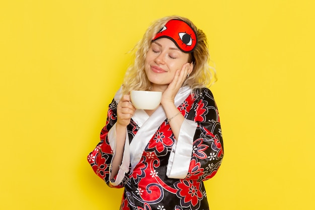 Vista frontal mujer joven en bata de noche y con máscara para los ojos preparándose para dormir tomando café sonriendo en la pared amarilla dormir modelo femenino cama de noche