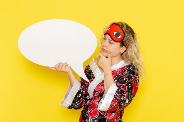 Vista frontal mujer joven en bata de noche y con máscara para los ojos sosteniendo un enorme cartel blanco en la pared de color amarillo claro, modelo de cama de noche para dormir, color de niña