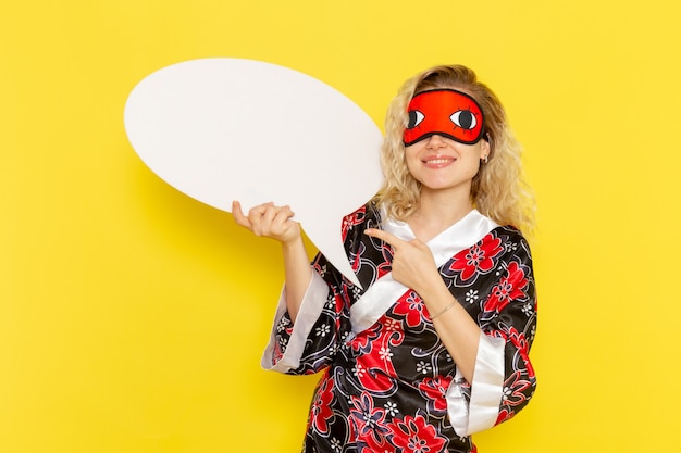 Vista frontal mujer joven en bata de noche sosteniendo un enorme cartel blanco y sonriendo en la pared amarilla dormir noche cama modelo niña color