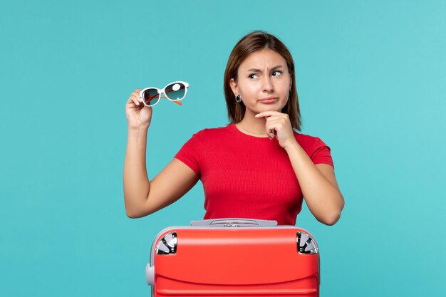 Vista frontal mujer joven con bolsa roja sosteniendo sus gafas de sol en el espacio azul claro