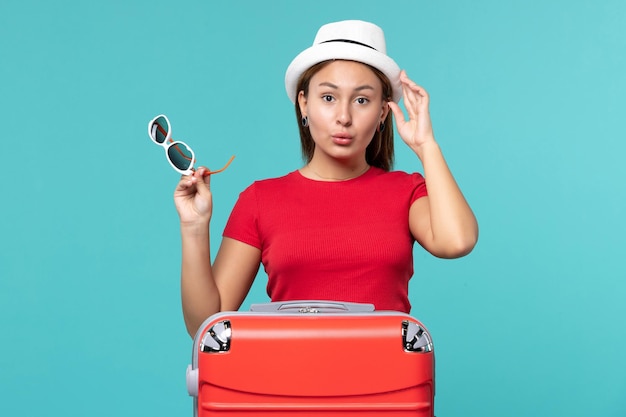 Vista frontal mujer joven con bolsa roja sosteniendo sus gafas de sol con sombrero en el espacio azul