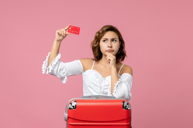 Vista frontal de la mujer joven con bolsa de vacaciones sosteniendo una tarjeta bancaria roja sobre el piso rosa modelo de mar viaje de vacaciones en color