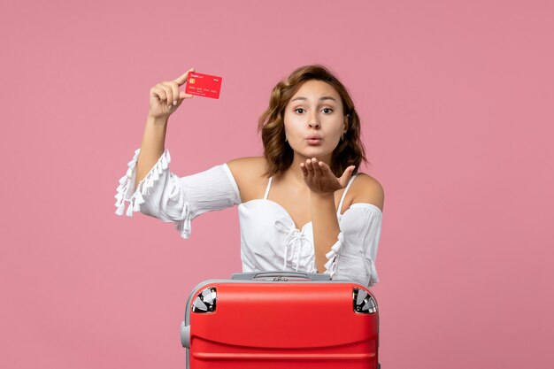 Vista frontal de la mujer joven con bolsa de vacaciones con tarjeta bancaria en la pared de color rosa claro