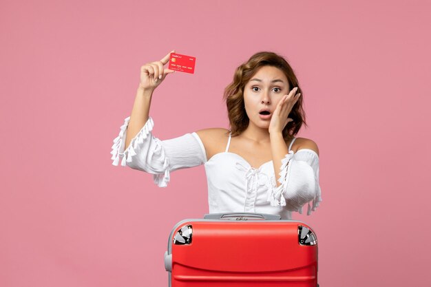 Vista frontal de la mujer joven con bolsa de vacaciones con tarjeta bancaria en la pared rosa