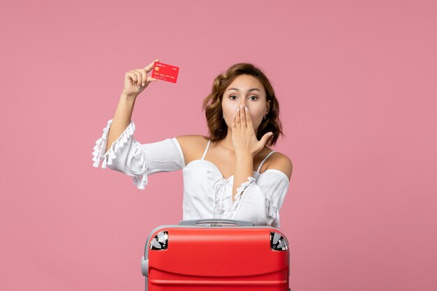 Vista frontal de la mujer joven con bolsa de vacaciones con tarjeta bancaria roja en la pared rosa