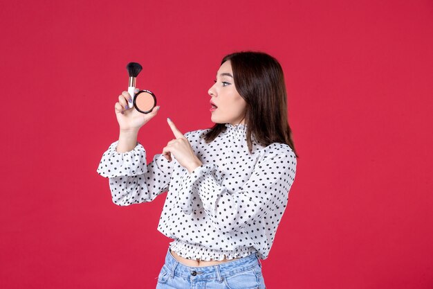 Vista frontal de la mujer joven con borla y polvo haciendo maquillaje en la pared roja