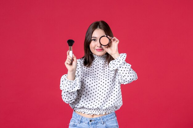 Vista frontal de la mujer joven con borla y polvo haciendo maquillaje en la pared roja