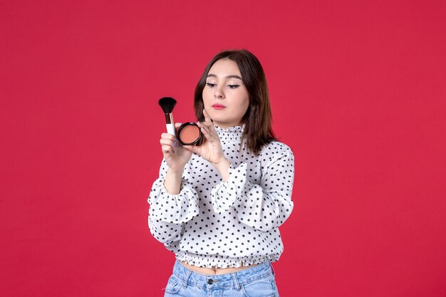 Vista frontal de la mujer joven con borla y polvo haciendo maquillaje en la pared roja