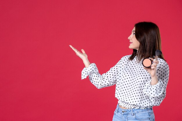 Vista frontal mujer joven con borla en polvo para maquillaje y pared roja