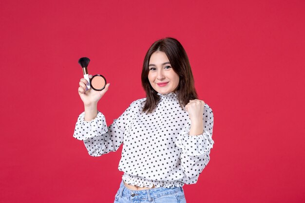 Vista frontal de la mujer joven con borla y polvo para maquillaje en pared roja