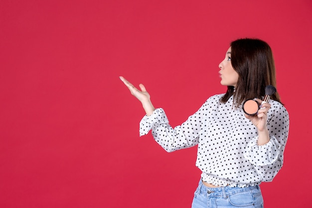 Vista frontal de la mujer joven con borla y polvo para maquillaje en pared roja