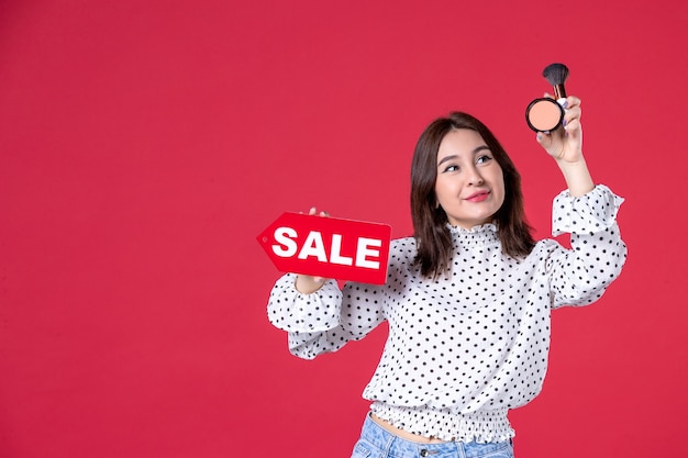 Vista frontal mujer joven con borla en polvo para maquillaje y placa de venta en pared roja