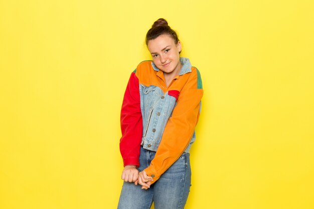 Una vista frontal mujer joven en camisa amarilla chaqueta colorida y jeans azul posando con una sonrisa en su rostro