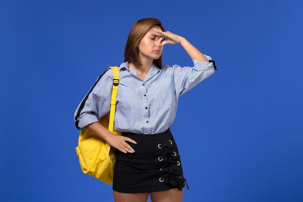 Vista frontal de la mujer joven en camisa azul falda negra con mochila amarilla cerrando la nariz en la pared azul claro