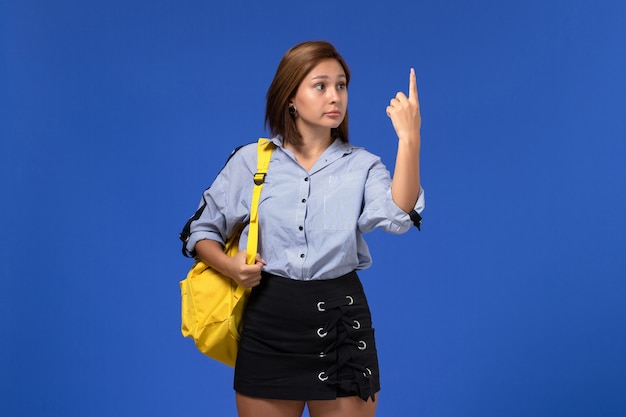 Vista frontal de la mujer joven en camisa azul falda negra con mochila amarilla levantando el dedo sobre la pared azul claro