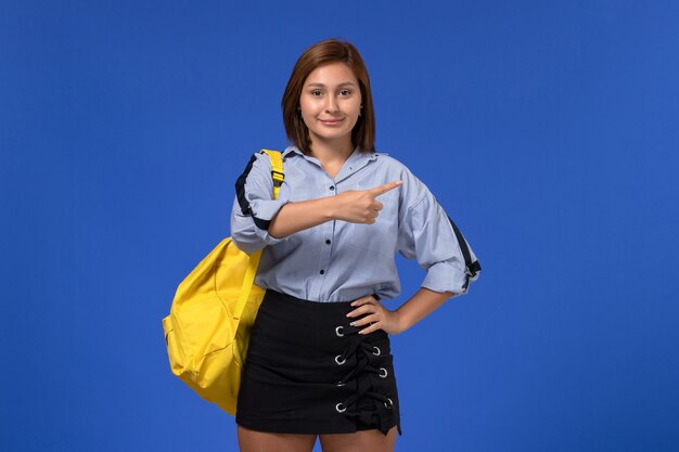 Vista frontal de la mujer joven en camisa azul falda negra con mochila amarilla posando sonriendo en la pared azul claro