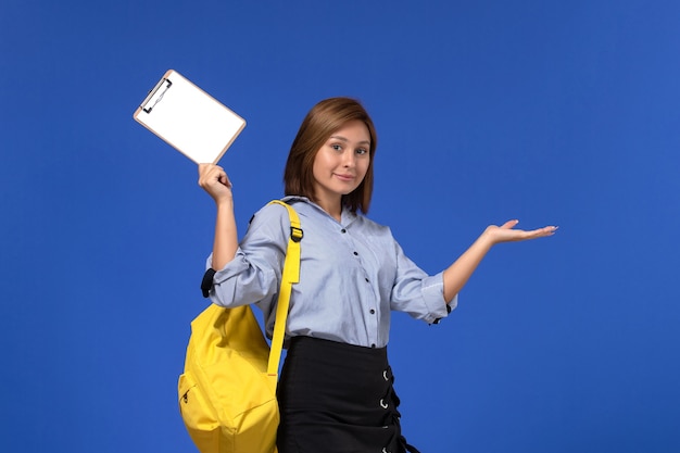 Vista frontal de la mujer joven en camisa azul falda negra con mochila amarilla sosteniendo el bloc de notas en la pared azul claro