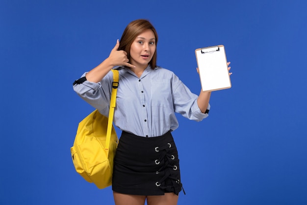 Vista frontal de la mujer joven en camisa azul falda negra con mochila amarilla sosteniendo el bloc de notas con una sonrisa en la pared azul