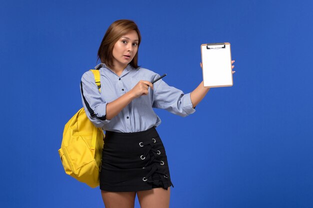 Vista frontal de la mujer joven en camisa azul falda negra con mochila amarilla y sosteniendo bolígrafo con bloc de notas en la pared azul