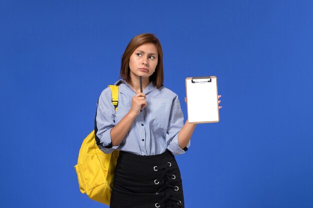 Vista frontal de la mujer joven en camisa azul falda negra con mochila amarilla y sosteniendo bolígrafo con bloc de notas en la pared azul