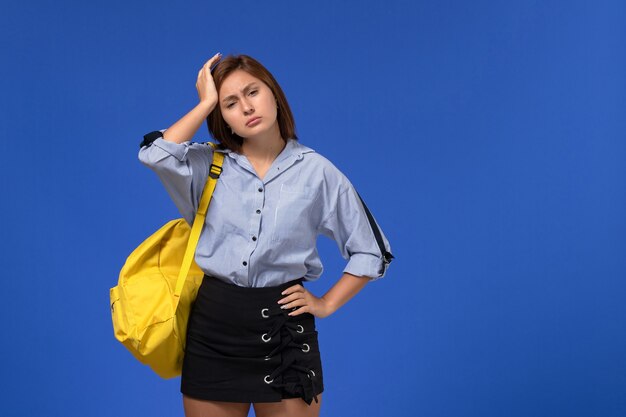 Vista frontal de la mujer joven en camisa azul con mochila amarilla con dolor de cabeza en la pared azul