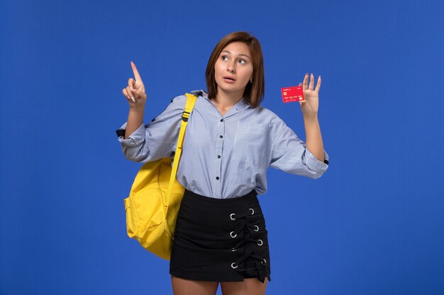 Vista frontal de la mujer joven en camisa azul con mochila amarilla sosteniendo una tarjeta de plástico roja en la pared azul