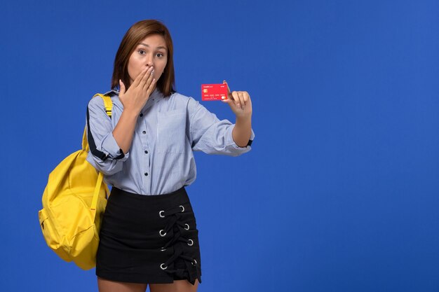 Vista frontal de la mujer joven en camisa azul con mochila amarilla sosteniendo una tarjeta de plástico roja en la pared azul