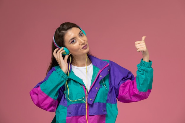 Vista frontal mujer joven con camisa blanca abrigo colorido escuchando música con una sonrisa en el escritorio de color rosa claro