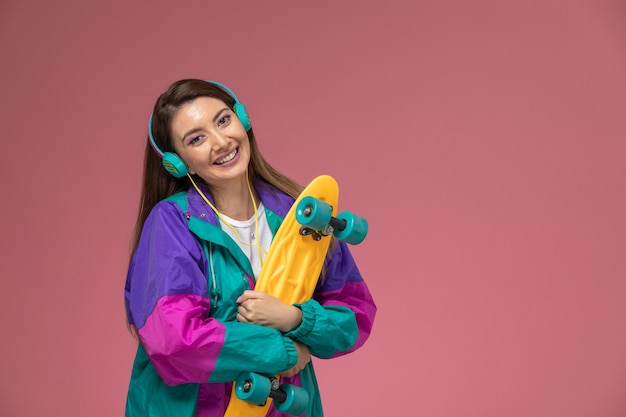 Vista frontal mujer joven con camisa blanca abrigo colorido escuchando música y sosteniendo patineta