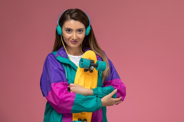 Vista frontal mujer joven con camisa blanca y abrigo colorido con patineta en la pared rosa, mujer modelo