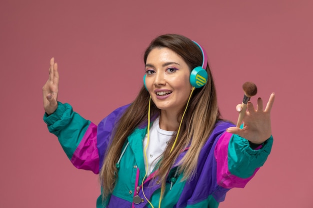 Vista frontal mujer joven en camisa blanca abrigo colorido sosteniendo pincel para maquillaje en pared rosa claro, pose de mujer modelo de mujer