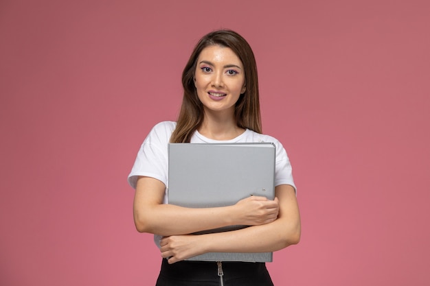 Vista frontal mujer joven en camisa blanca con archivo gris con sonrisa en la pared rosa, mujer de color pose modelo mujer