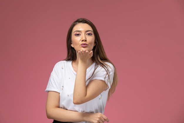 Vista frontal mujer joven en camisa blanca enviando besos al aire en la pared rosa, modelo de mujer de color posando mujer