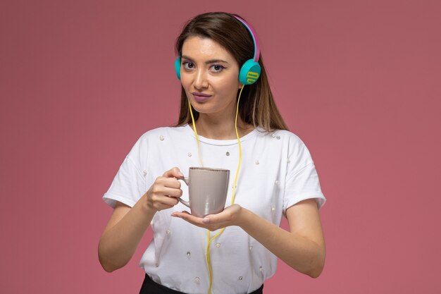 Vista frontal mujer joven en camisa blanca escuchando música con policía con café en la pared rosa, mujer de color modelo mujer