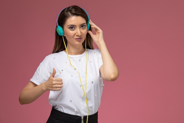 Vista frontal mujer joven con camisa blanca escuchando música a través de auriculares en la pared rosa, mujer de color pose modelo mujer