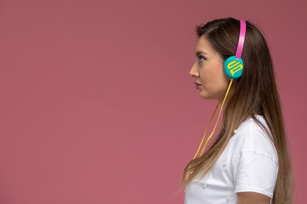 Vista frontal mujer joven con camisa blanca escuchando música a través de sus auriculares en la pared rosa, mujer de color pose modelo mujer