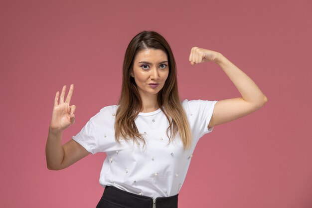 Vista frontal mujer joven en camisa blanca flexionando en modelo de pose de mujer de color de pared rosa