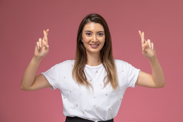 Vista frontal mujer joven en camisa blanca mostrando los dedos cruzados sonriendo en la pared rosa, color mujer modelo mujer