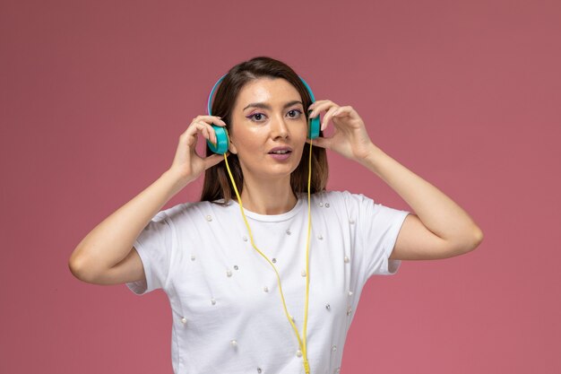 Vista frontal mujer joven con camisa blanca posando y escuchando música a través de sus auriculares en la pared rosa, modelo de mujer de color posando mujer