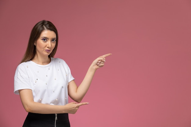 Vista frontal mujer joven en camisa blanca posando con la mano levantada en la pared rosa, modelo de mujer de color posando mujer