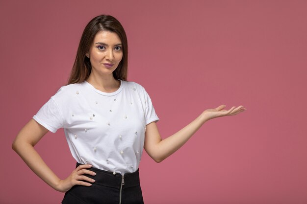 Vista frontal mujer joven en camisa blanca posando con la mano levantada en la pared rosa, modelo mujer pose mujer