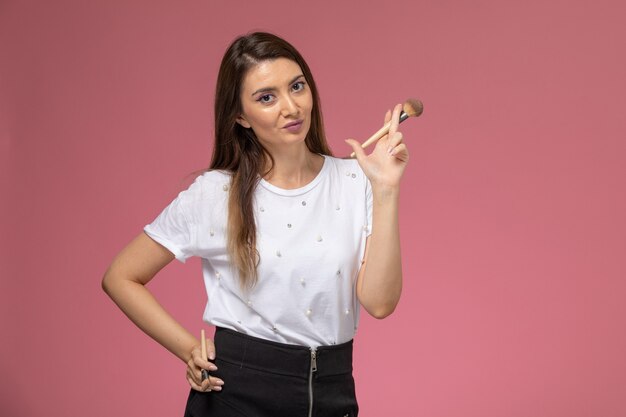 Vista frontal mujer joven en camisa blanca posando con pincel de maquillaje en pared rosa, modelo mujer pose mujer