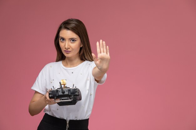 Vista frontal mujer joven con camisa blanca que muestra la señal de stop con control remoto en la pared rosa, modelo mujer pose mujer