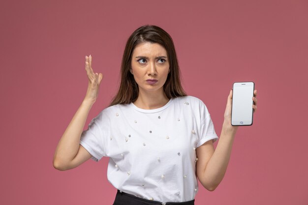 Vista frontal mujer joven en camisa blanca con smartphone en pared rosa claro, pose de mujer modelo mujer