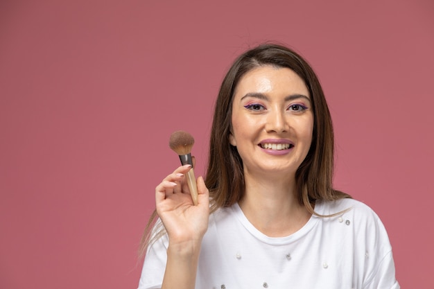 Vista frontal mujer joven en camisa blanca sonriendo y sosteniendo el pincel de maquillaje en la pared rosa