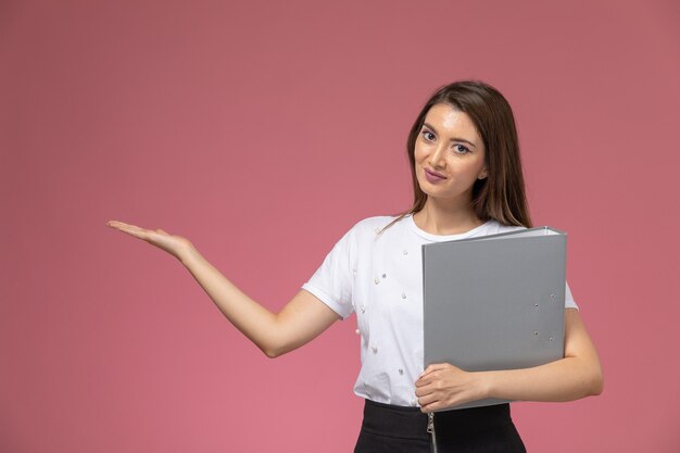 Vista frontal mujer joven con camisa blanca sosteniendo archivos grises en la pared rosa claro, pose de mujer modelo