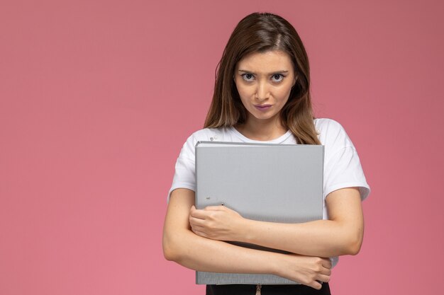 Vista frontal mujer joven con camisa blanca sosteniendo archivos grises en la pared rosa claro, pose de mujer modelo